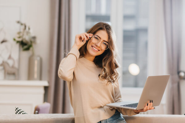 Cute girl in beige shirt touching glasses and holding laptop with smile. Indoor photo of beautiful female student preparing for lesson with computer at home..